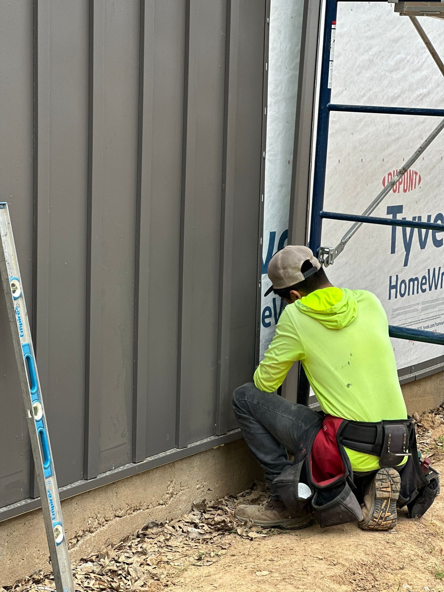 Worker in neon jacket kneeling by wall, installing siding beside a window on a construction site.