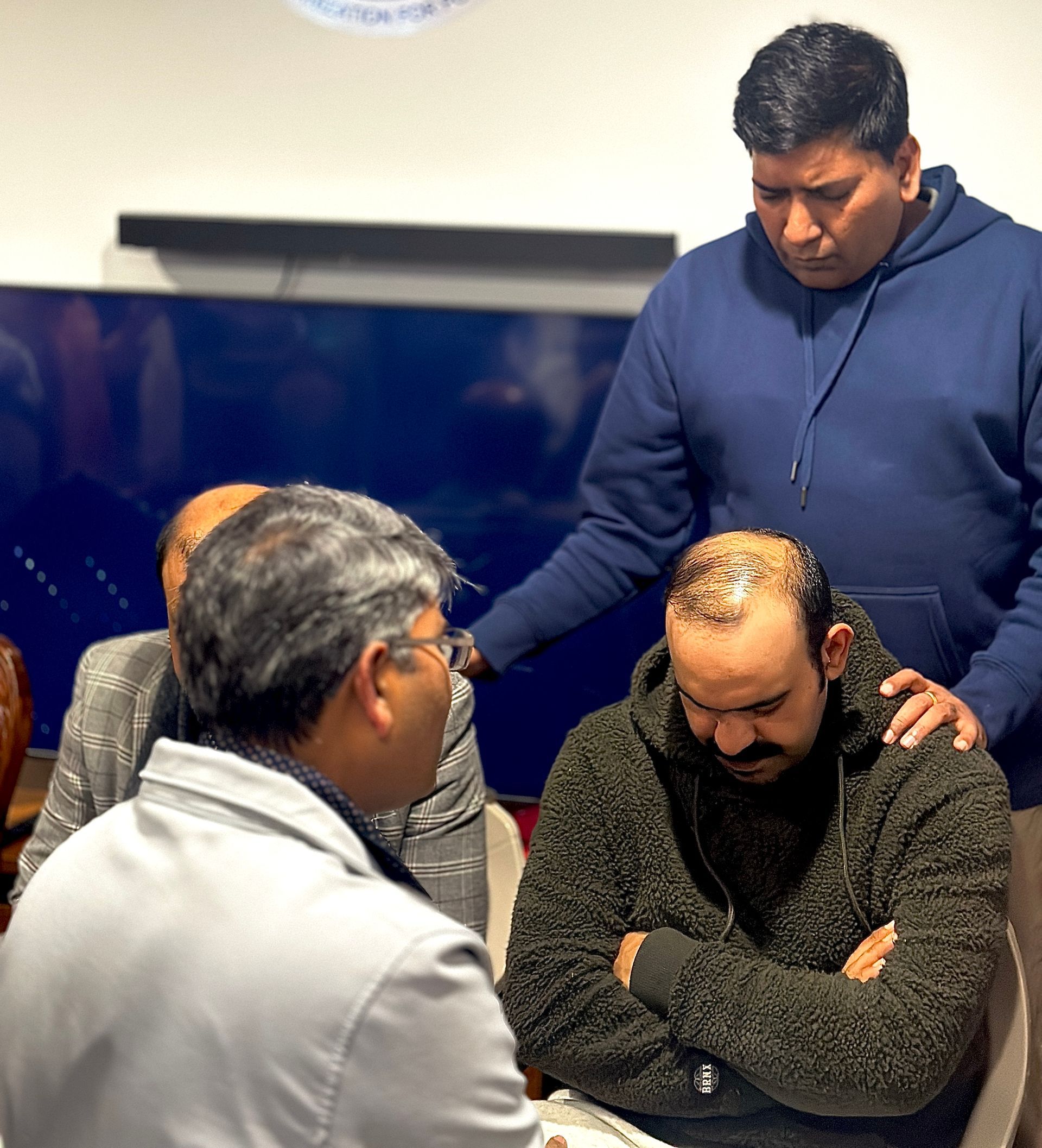 Man with hand on chest, surrounded by others, inside a building with an arched window.