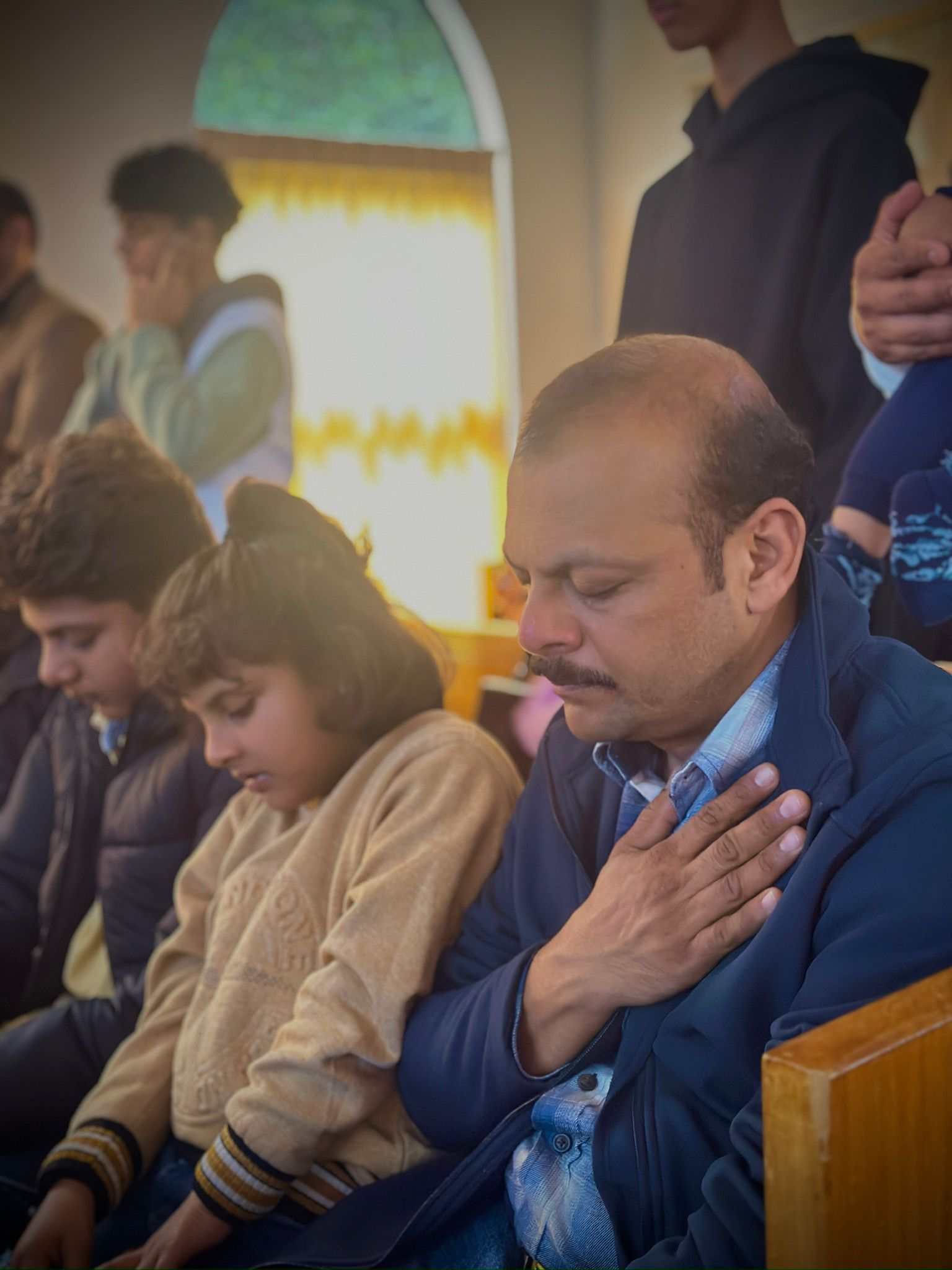 Man with hand on chest, surrounded by others, inside a building with an arched window.