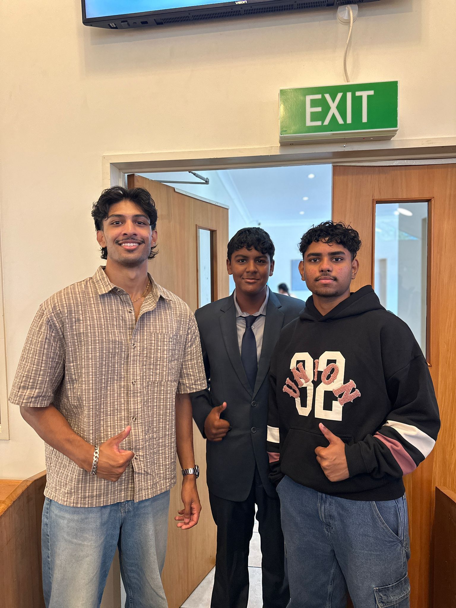 Three young people pose inside near an exit sign. Two give thumbs up, smiles present.