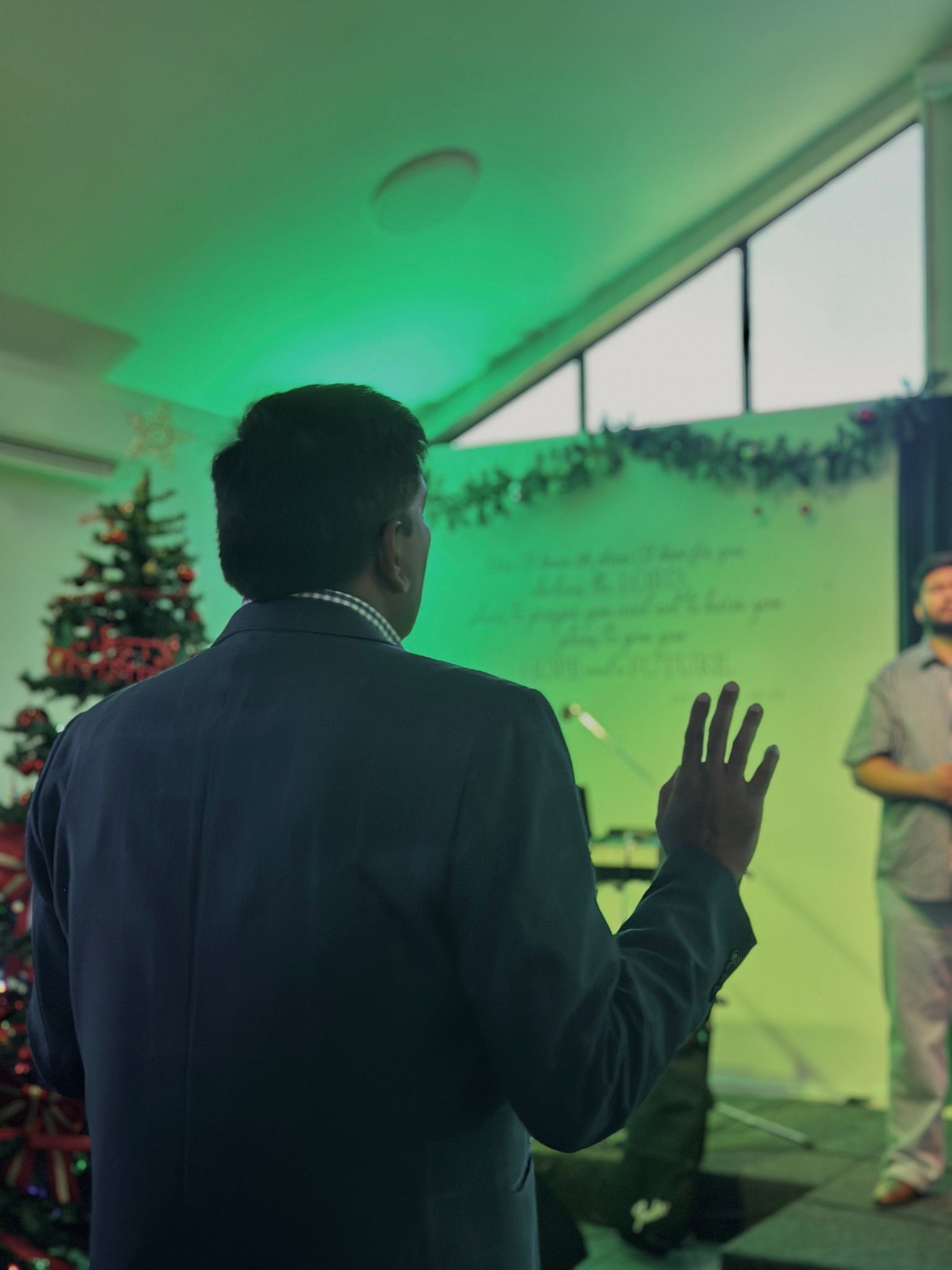 Man in suit speaking at podium, gesturing. Green-lit stage with Christmas tree and background text visible.