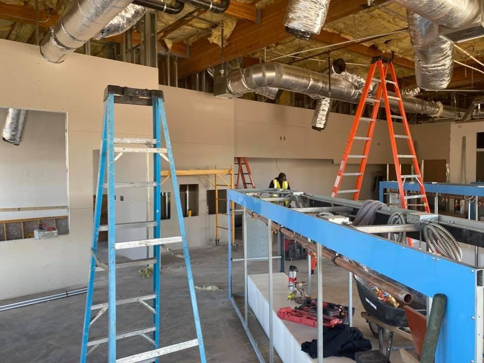 Construction workers inside an unfinished building with ladders, exposed ceiling pipes, and metal framing structures.