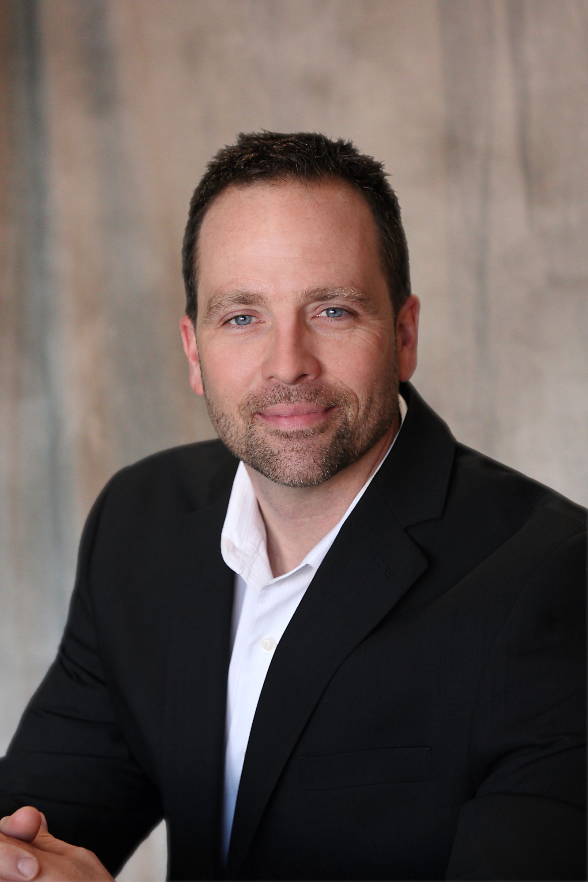 A man in a black blazer and white shirt, looking forward with a neutral expression against a grey background.