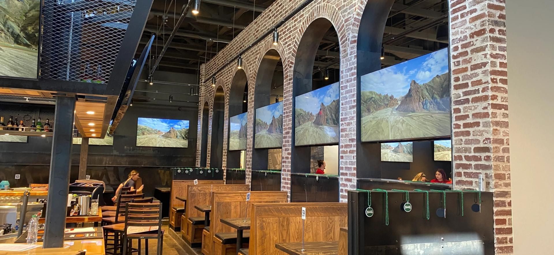 A row of booths in a restaurant featuring brick arches, wood furniture, and TVs mounted within each archway.