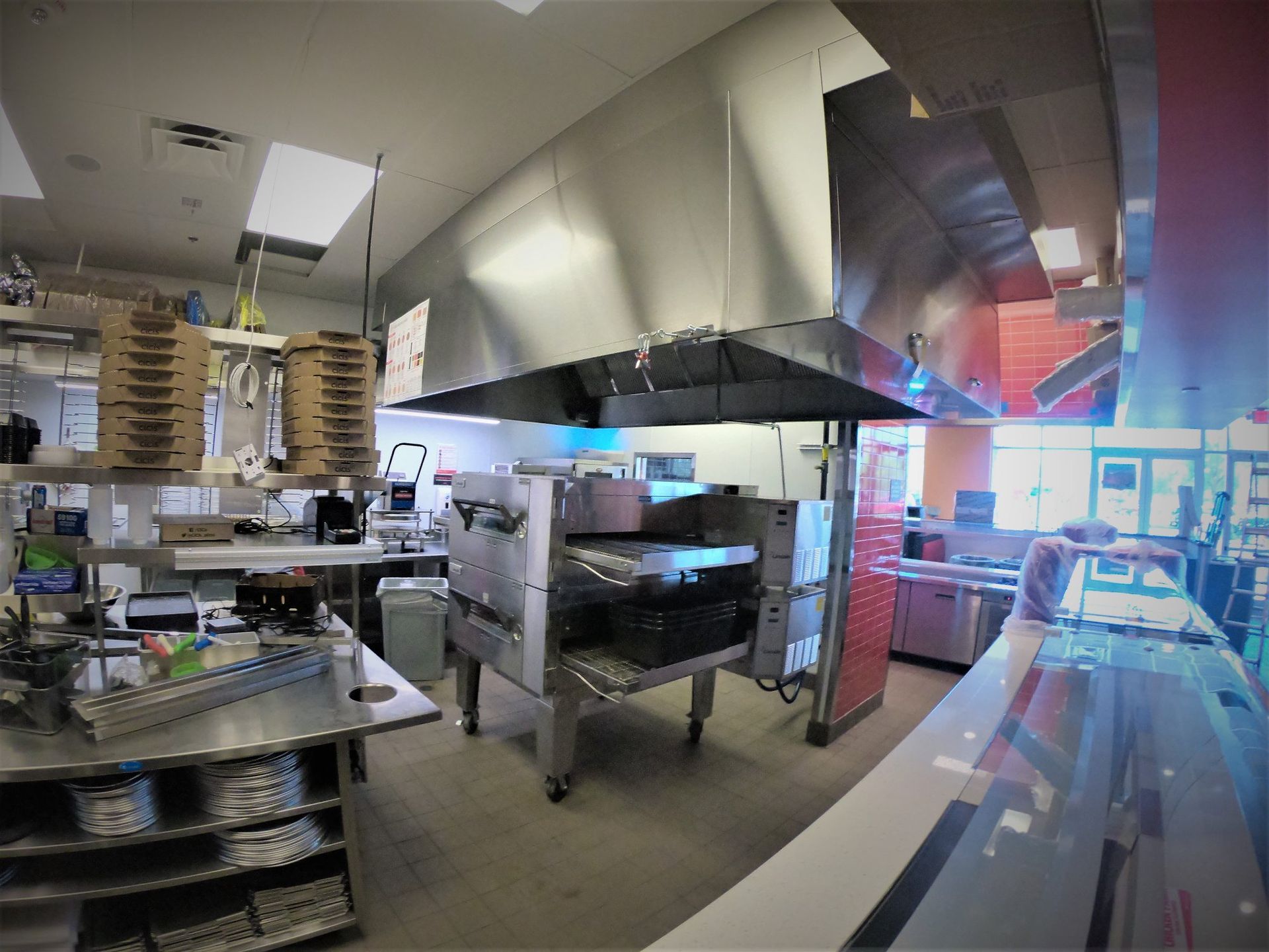 A commercial kitchen showing a stainless steel pizza oven under a large ventilation hood and stacks of pizza boxes.