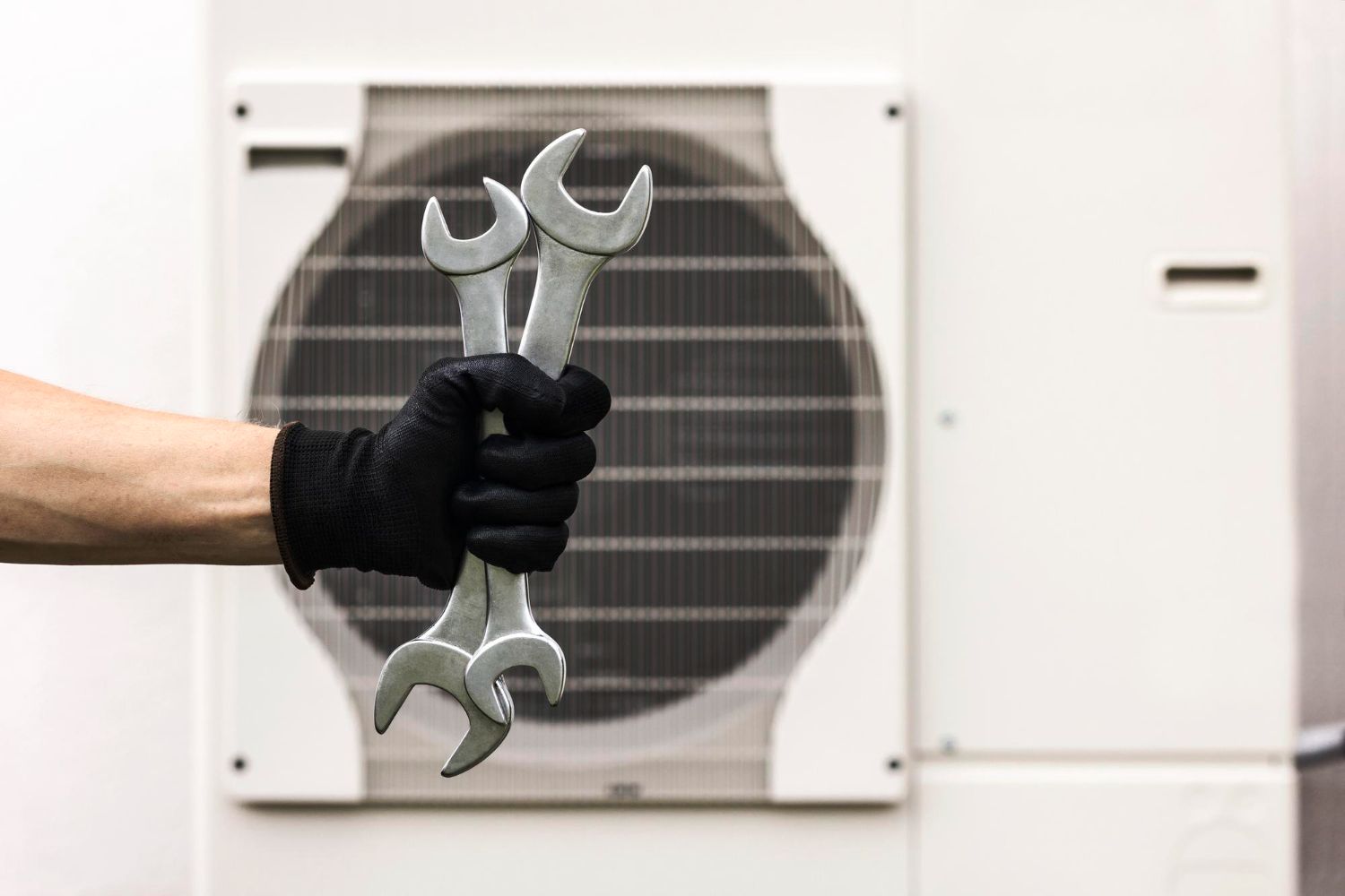 Hand in black glove holding two wrenches in front of an AC unit.