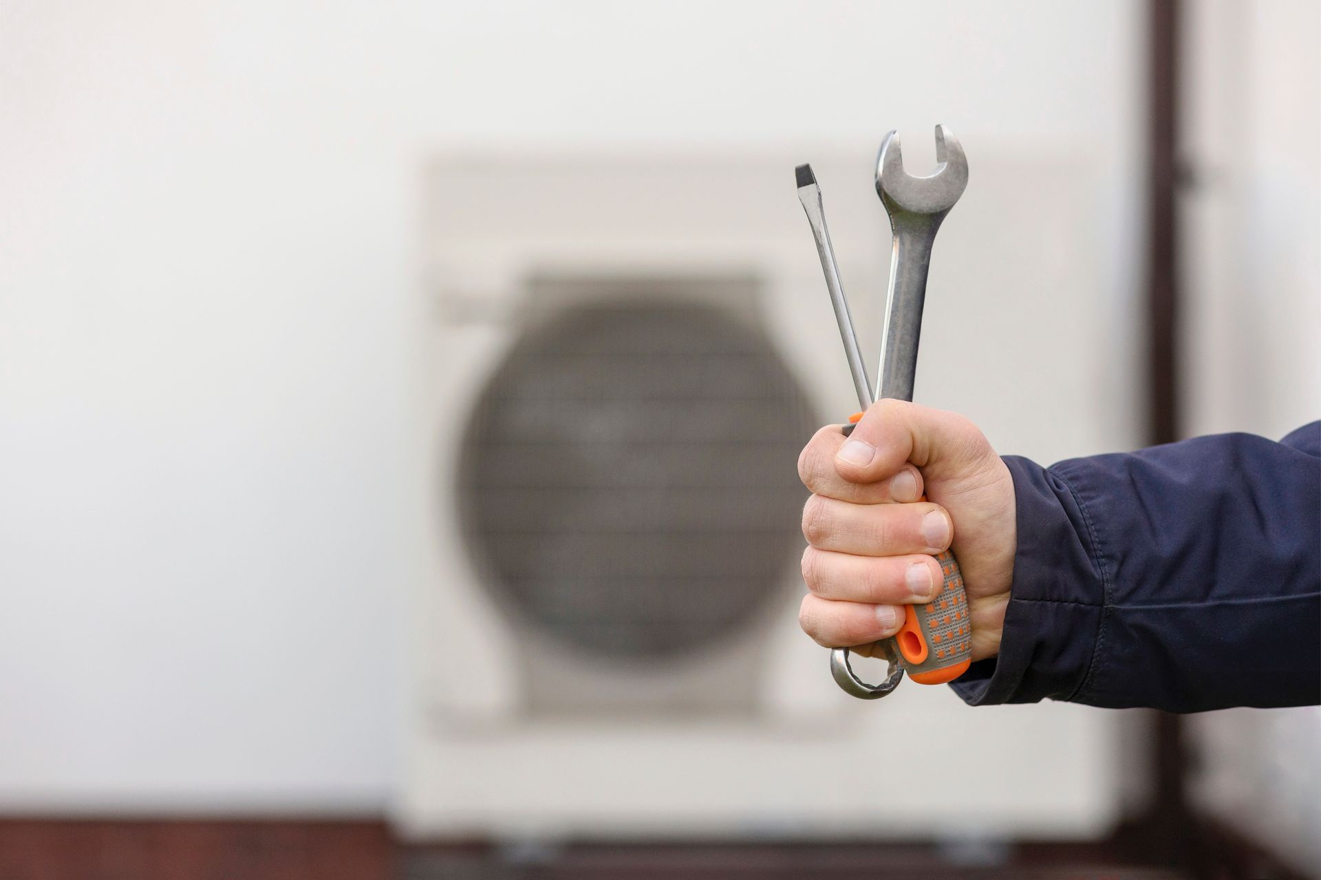Hand holding two wrenches in front of an outdoor air conditioner unit