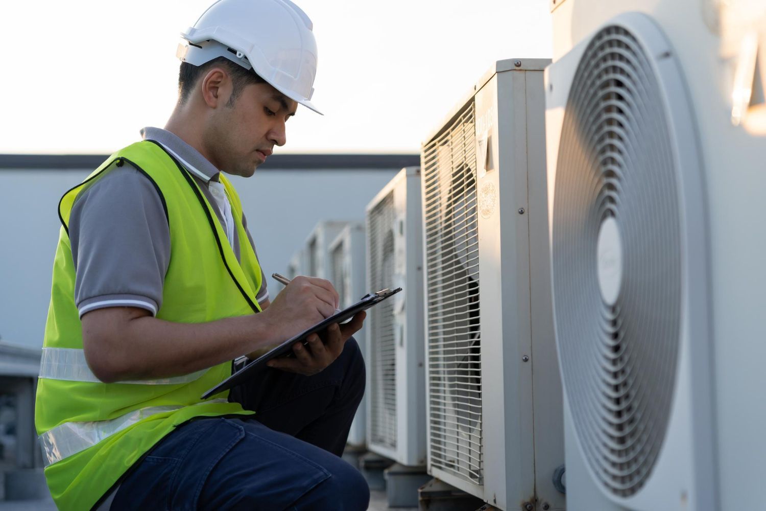 A man is kneeling down and writing on a clipboard in front of air conditioners.