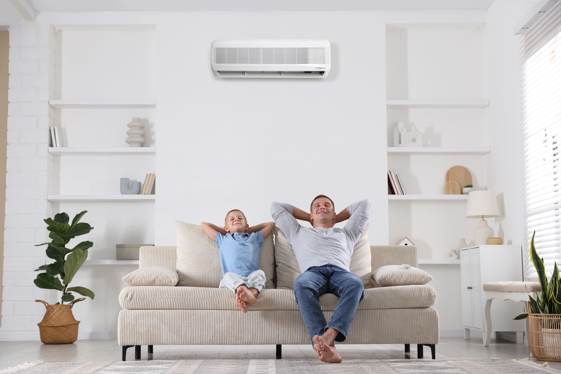Two people relaxing on a couch in a bright living room with white shelves and a wall-mounted air conditioner.