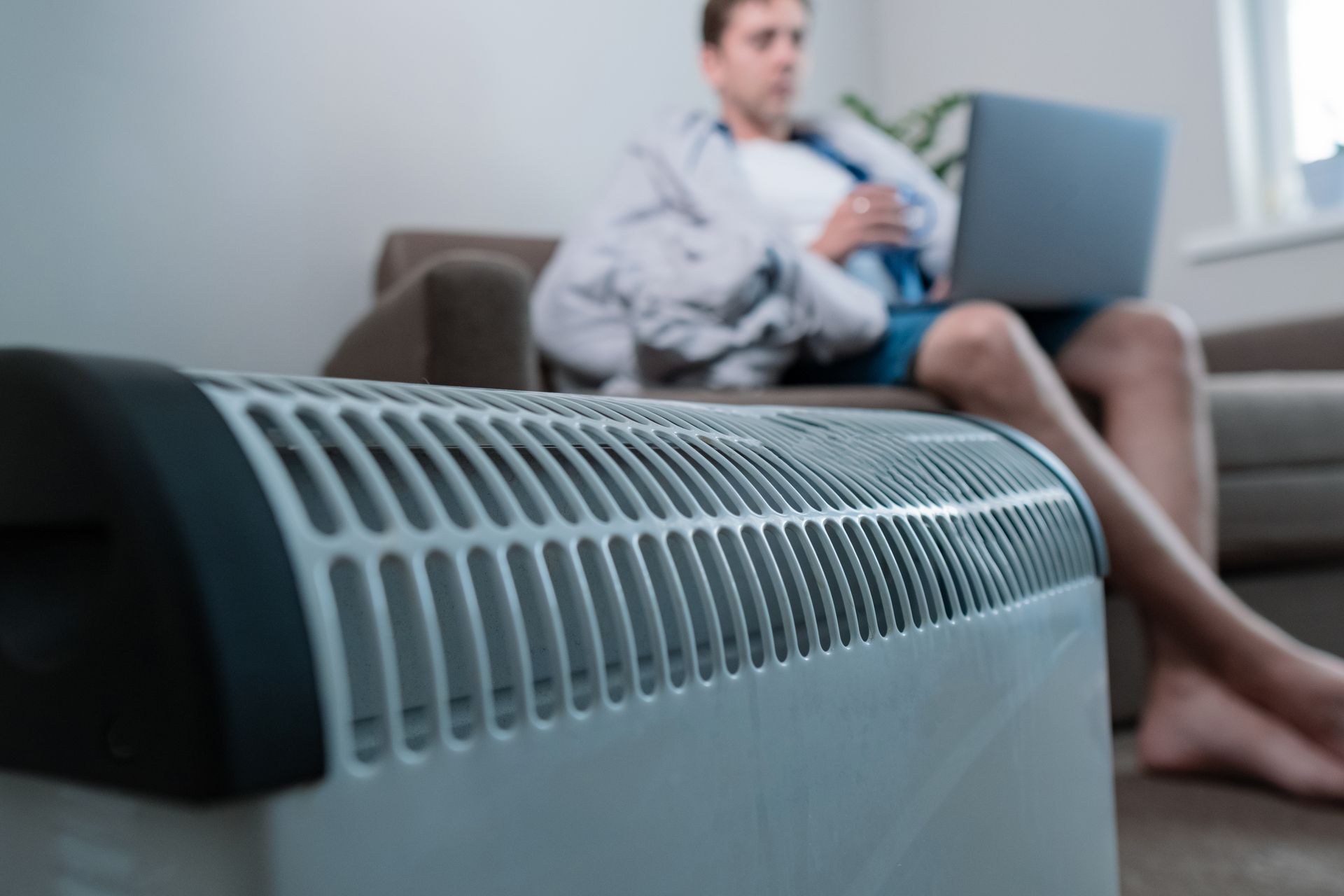 A man is sitting on a couch with a laptop and a heater in the foreground.