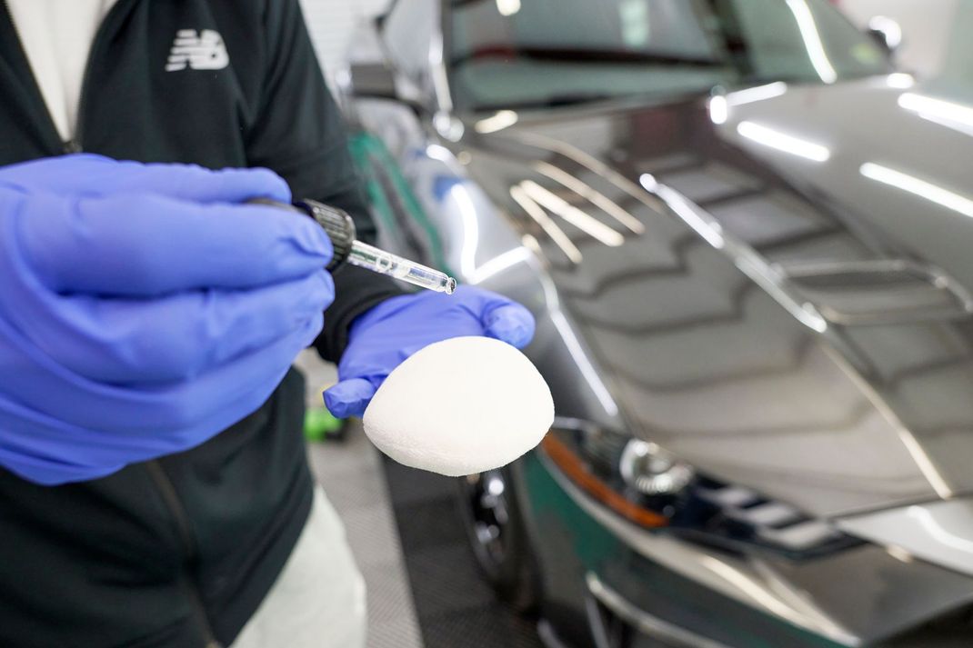 A person wearing blue gloves is applying ceramic coating to an application pad in front of a car.