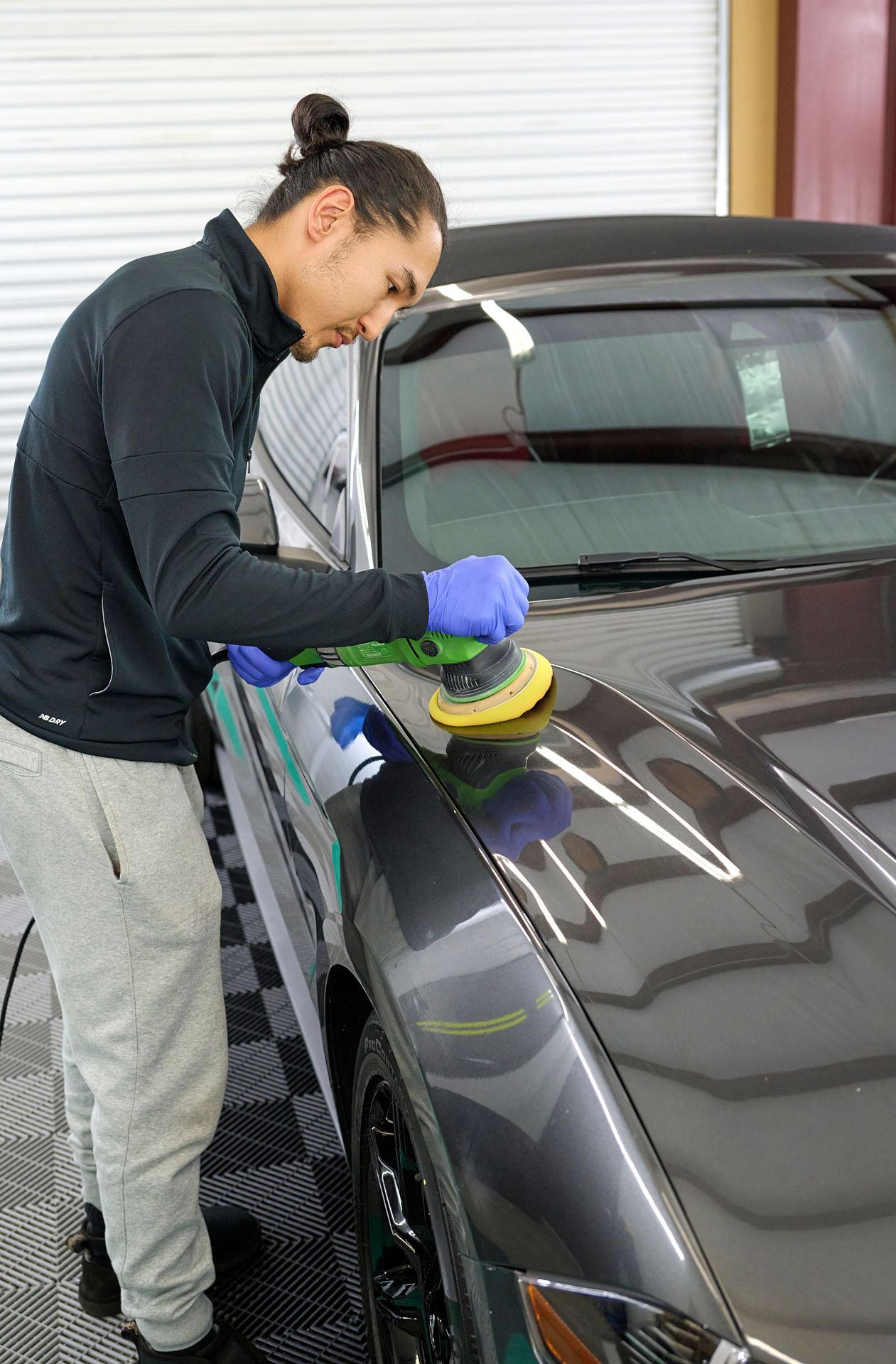 A professional technician, Max, owner and operator of GT Tinting, carefully applying a ceramic coating to a sleek black car using a dual-action polisher. He is wearing a black long-sleeve shirt, gray sweatpants, gloves, and standing in a clean workshop.