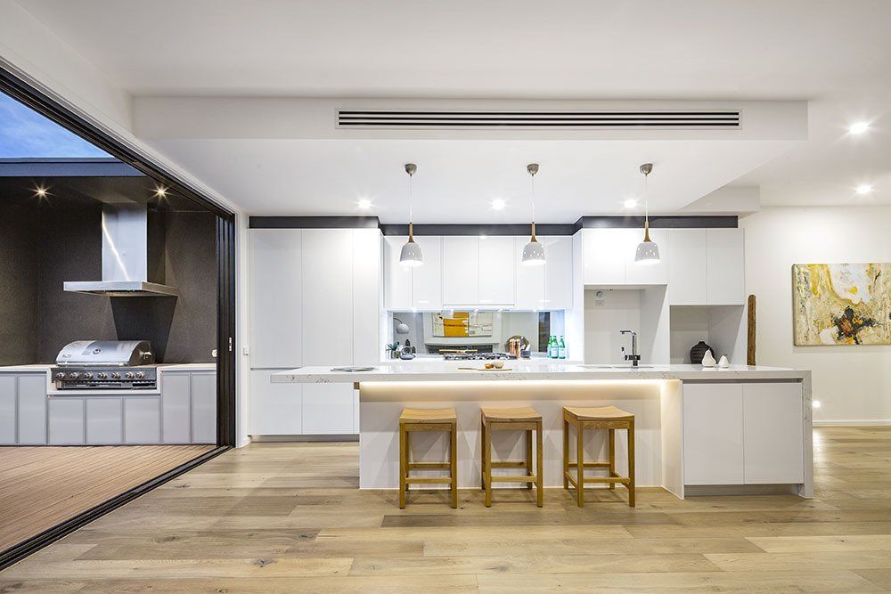 A kitchen with white cabinets , wooden floors , stools and a grill.