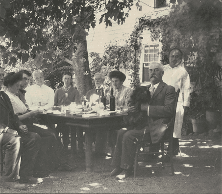 A family sits around a dining table outdoors, with Ah Hing dressed in a white shirt and apron standing in the background.