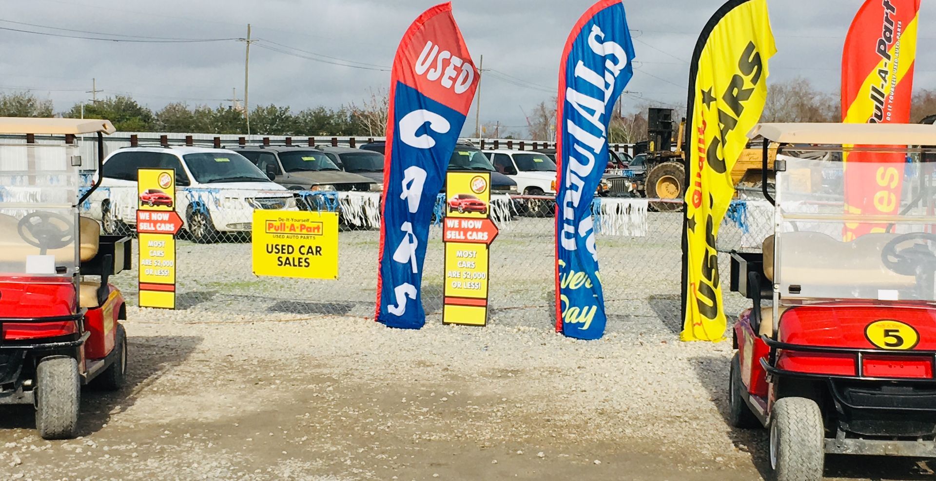 Used car lot entrance with golf carts, flags, and cars in the background on a cloudy day.