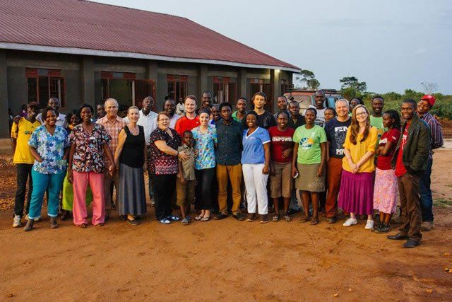 Group of people posing in front of a building. Diverse faces smiling, outdoors, some wearing colorful clothes.