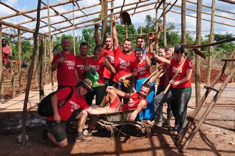 Group of people in red shirts inside a wooden structure frame, celebrating. Some hold tools, arms raised, happy expressions.