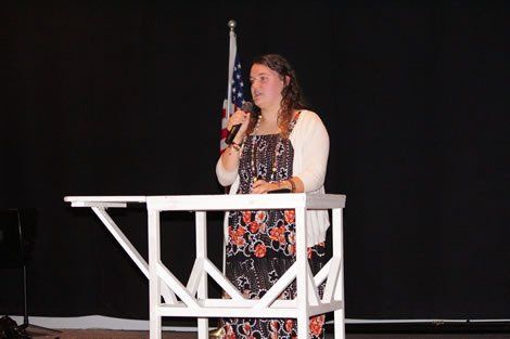 Woman speaking at a podium, American flag in background, dark setting.