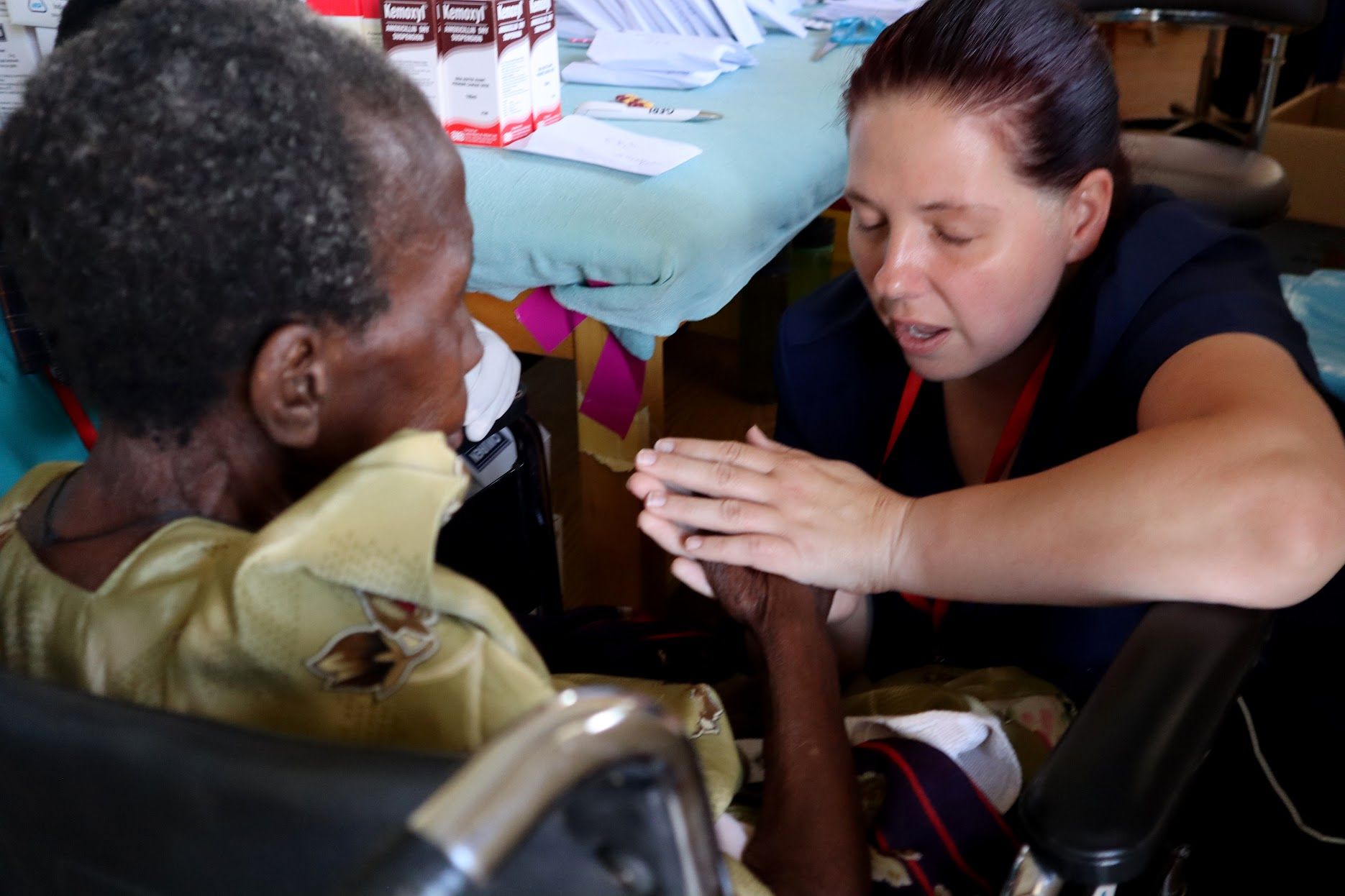 A healthcare worker comforts an elderly person in a wheelchair, holding hands.
