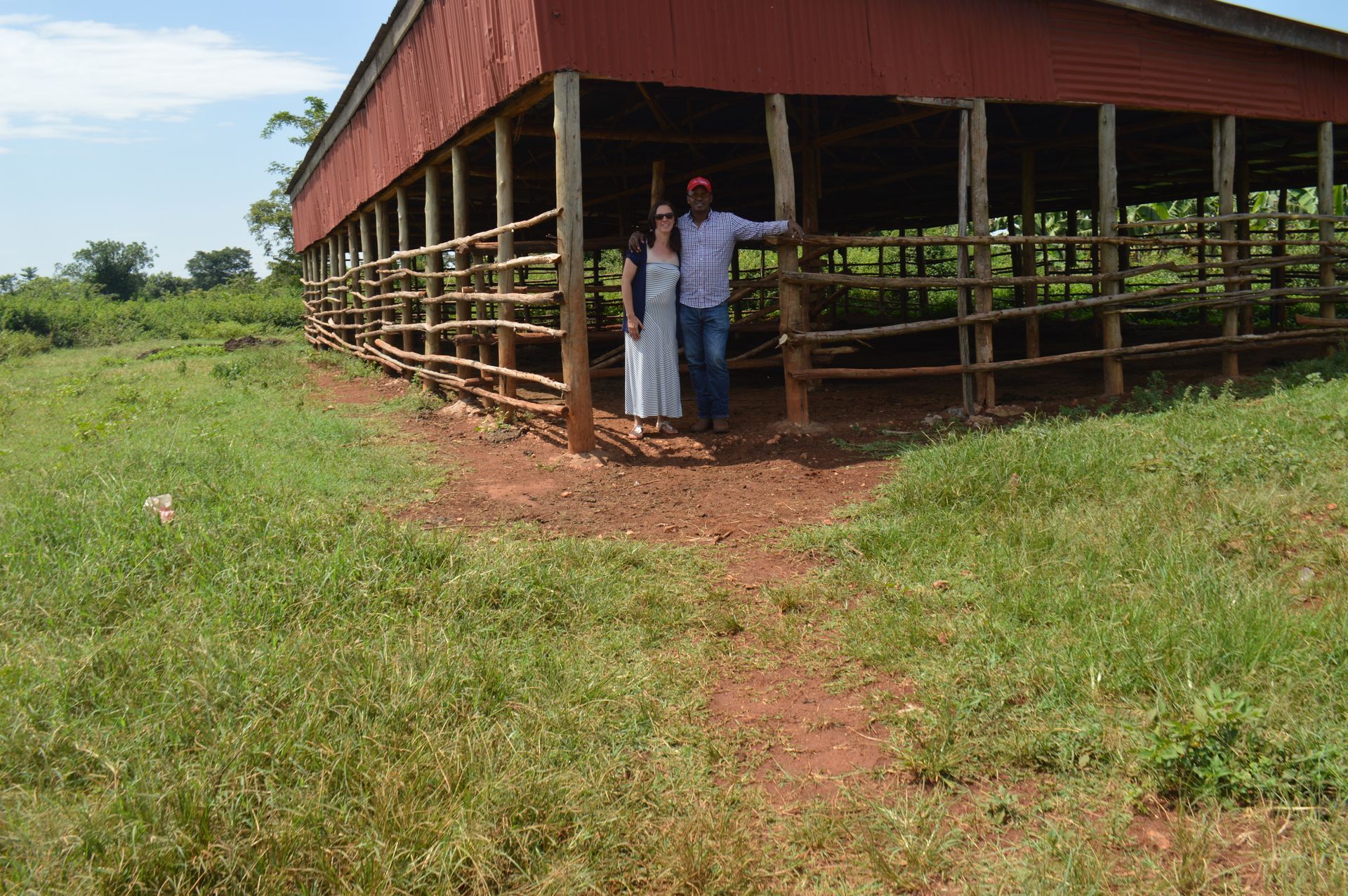 Couple stands under a red barn's open side, surrounded by a grassy field.