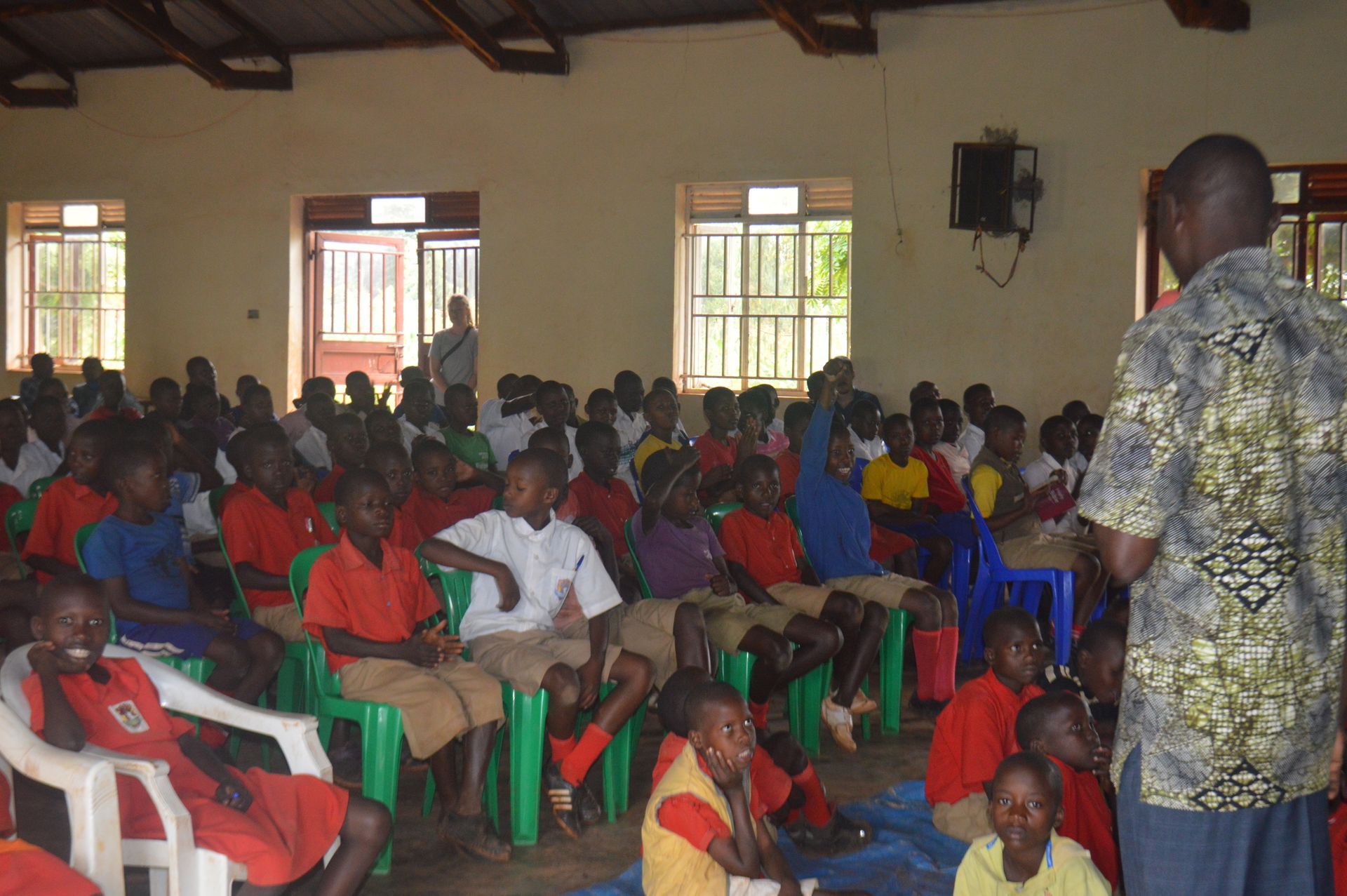 A man speaks to a group of children in a room. Children are seated, wearing red and blue uniforms.