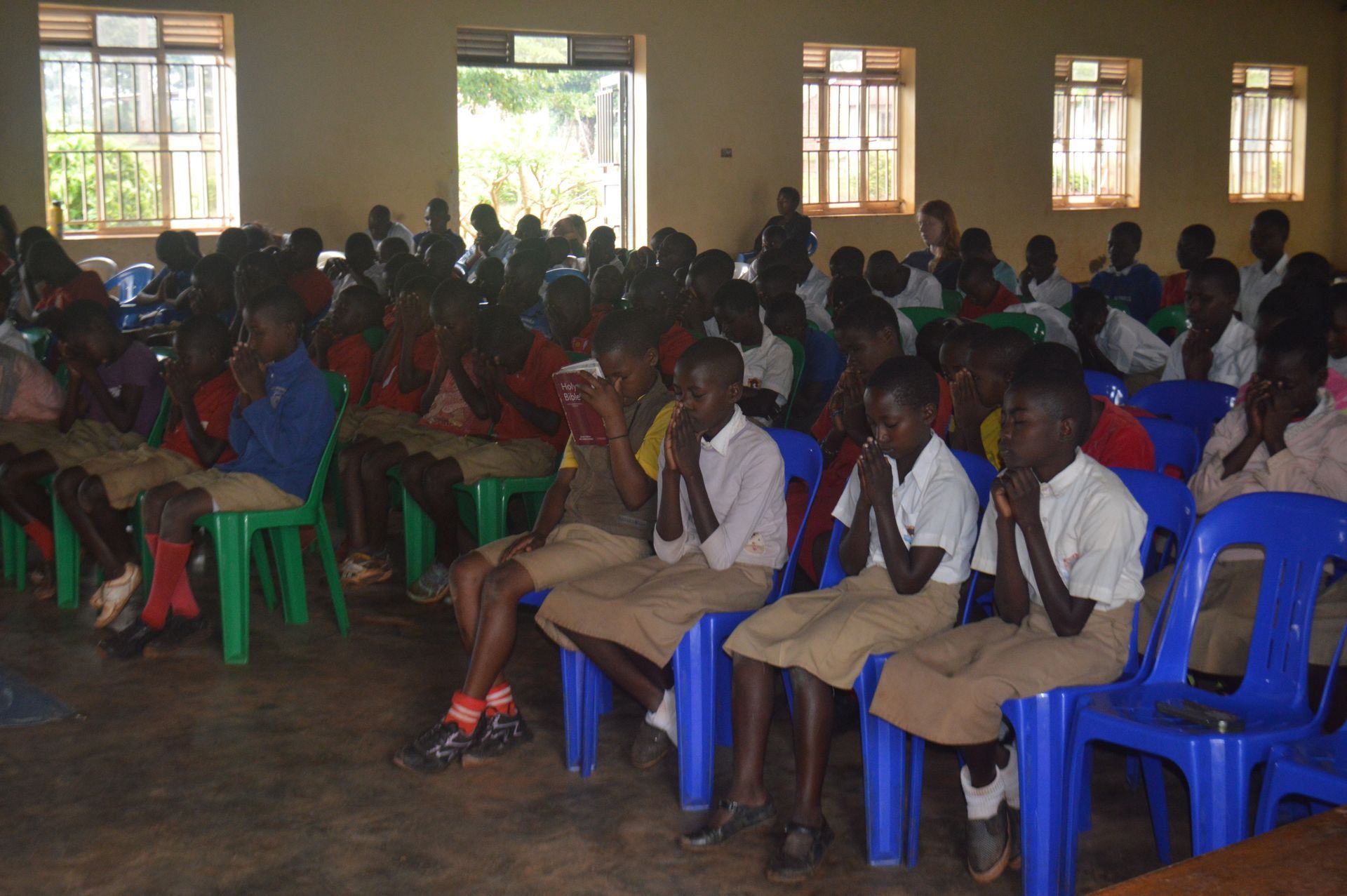 Children in school uniforms seated in a room, hands clasped. Many faces look forward; some wear blue or green chairs.