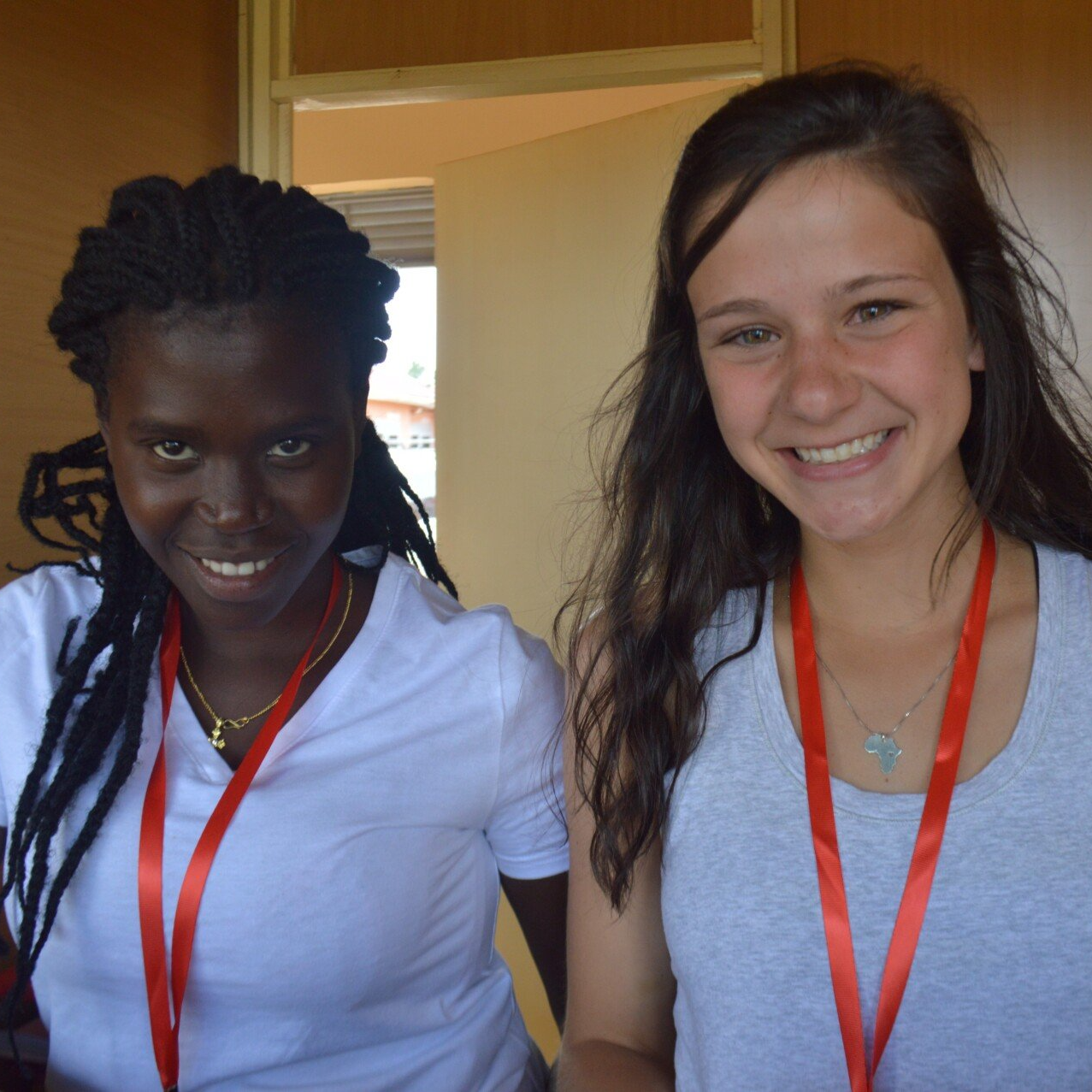 Two young women smiling, one Black with braids, one white with long hair, wearing red lanyards.