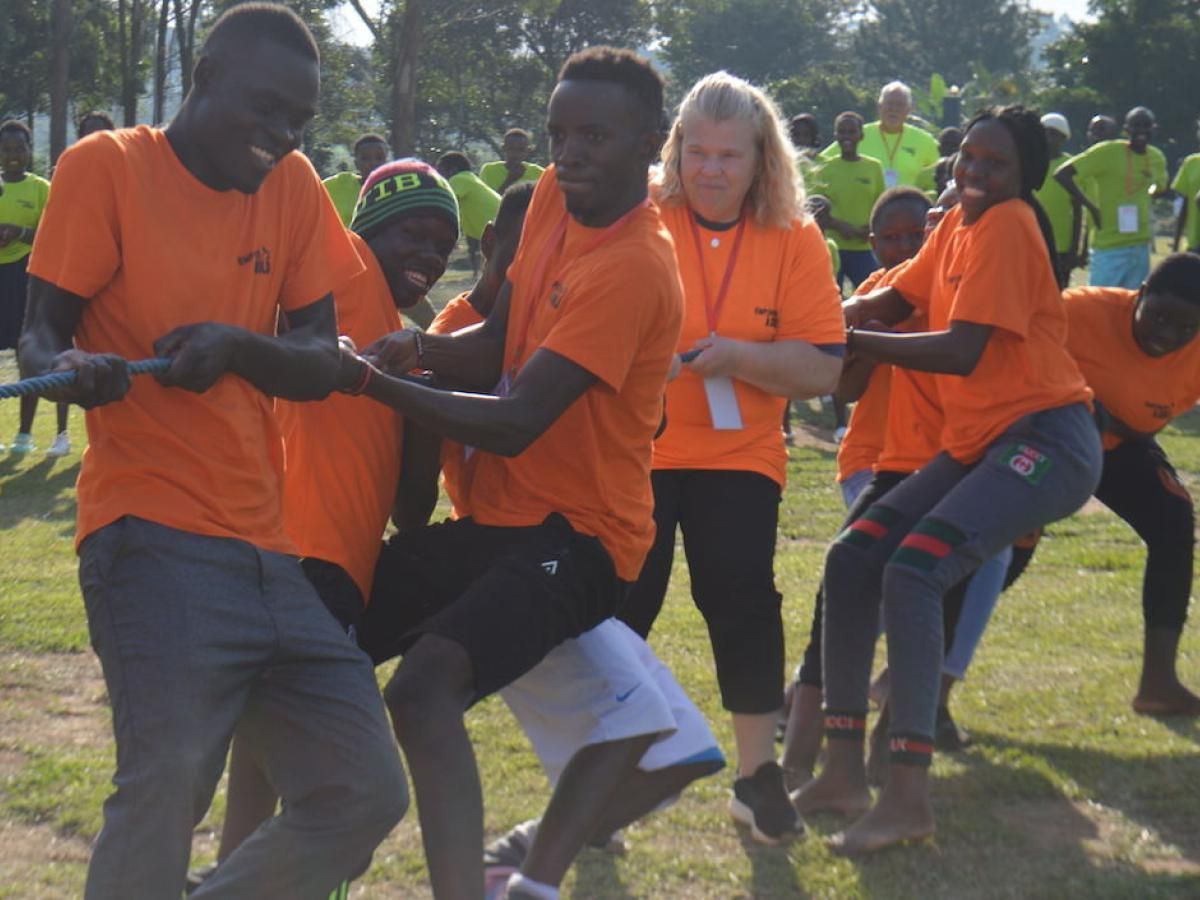 People in orange shirts play tug-of-war on grass; a woman watches.