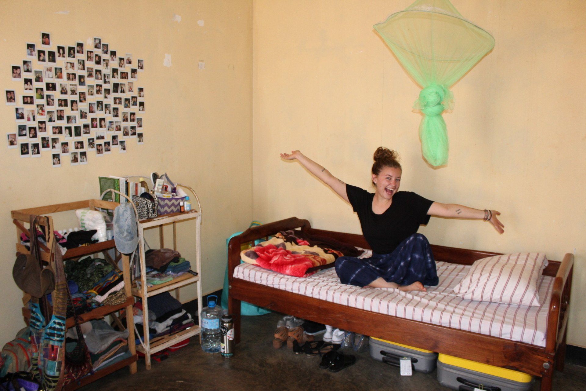 Woman in a room with arms outstretched, sitting on a bed. Wall with photos and shelves.