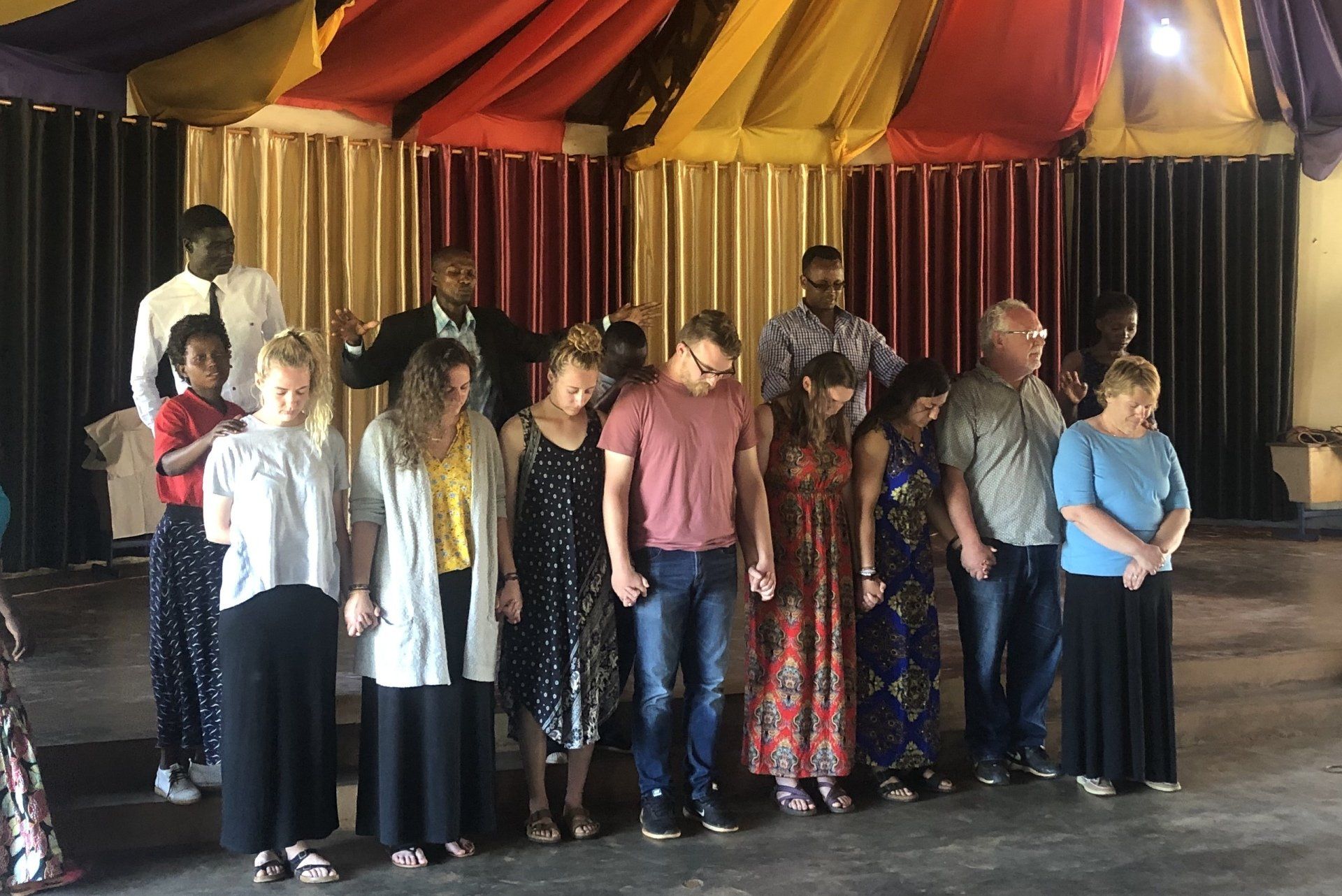 Group of people, eyes closed, hands clasped, in a ceremony. Colorful curtains in the background.