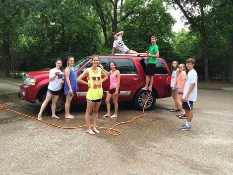 Group of teens washing a red SUV outdoors on a cloudy day. Some are standing on the vehicle.