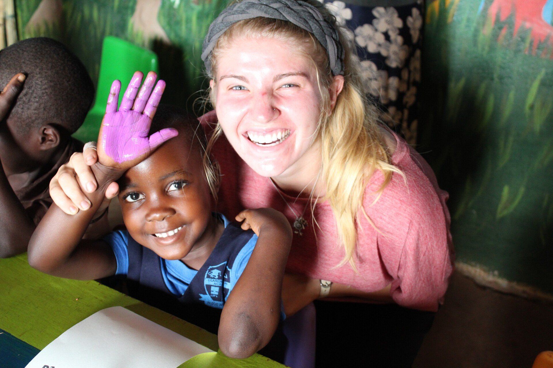 Woman smiles with child, holding up a hand painted purple, seated at a table indoors.