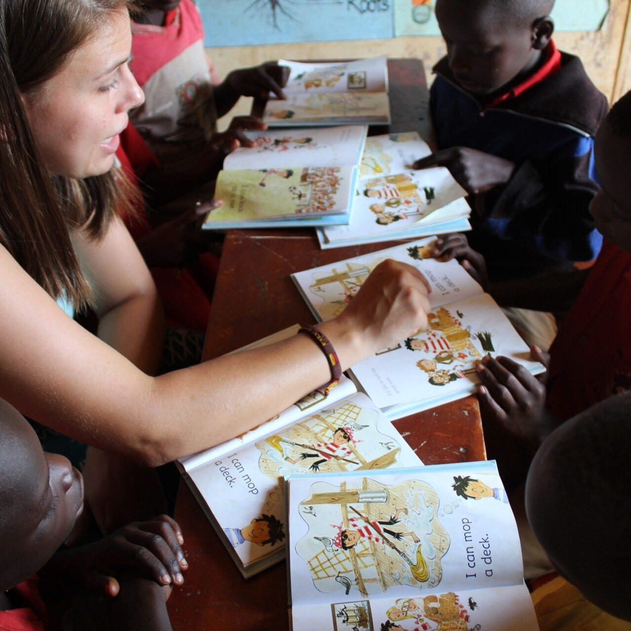 Woman teaching children, pointing at a book. They sit around a table, reading and smiling.