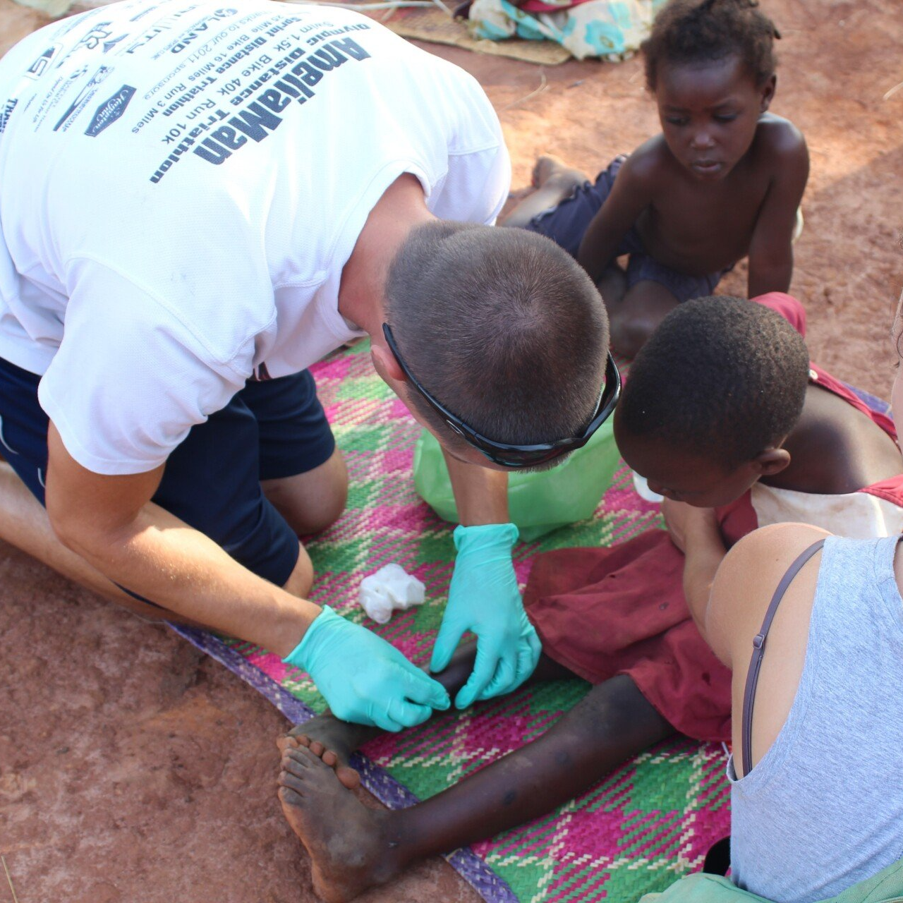 Man tending to a child's foot wound on a mat; another child watches.