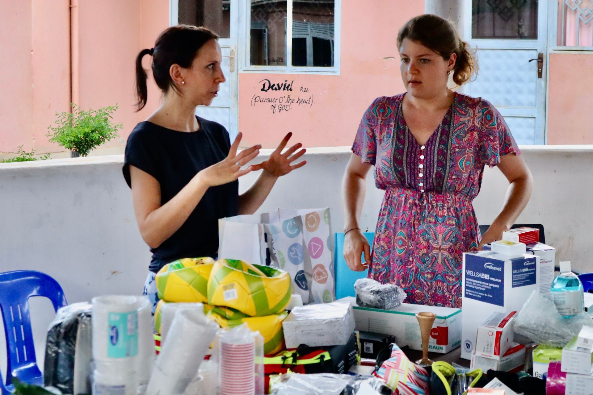 Two women at a table with supplies, one gesturing, likely distributing aid. Outdoors, sunny.