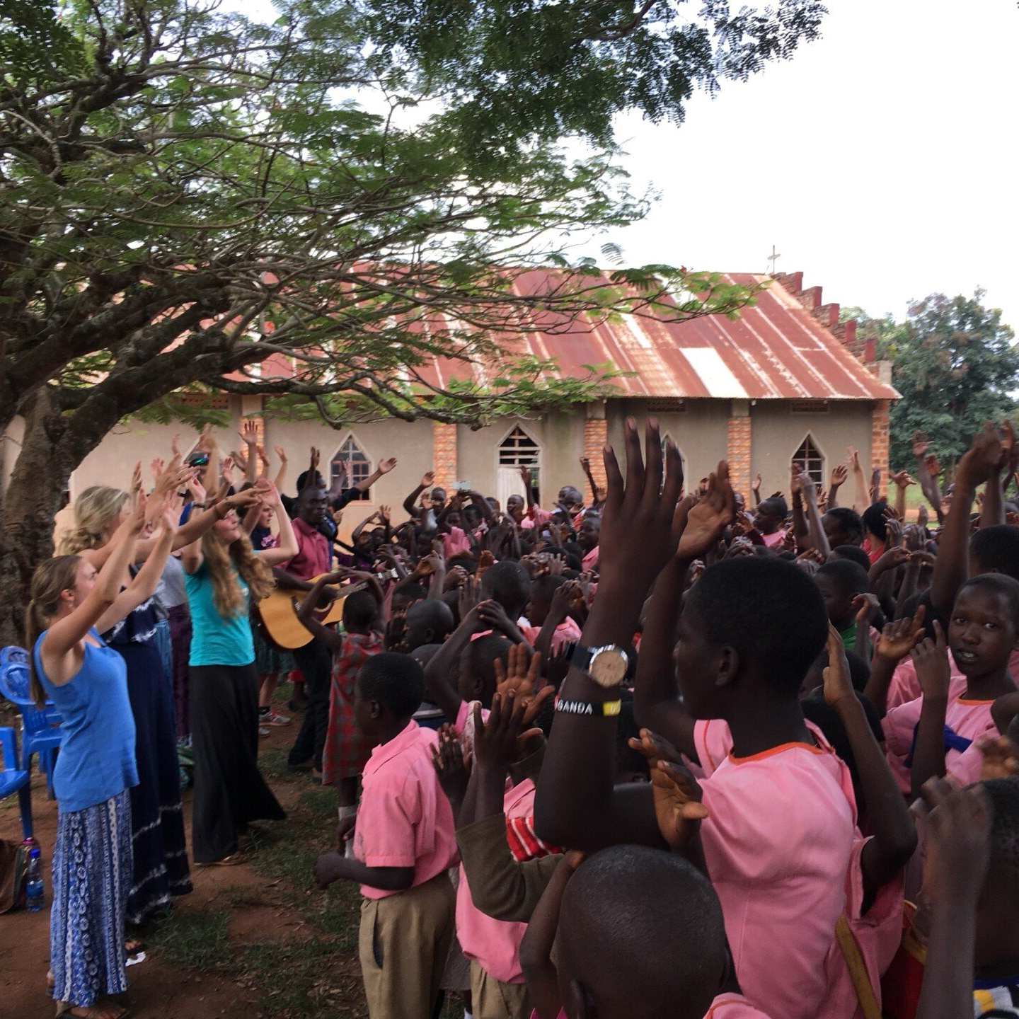 People with raised arms sing and dance outside a building with a tin roof. Some play music.