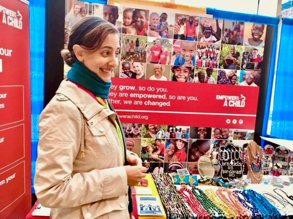 Woman at a booth, smiling, looking at jewelry. A poster with photos of children and text 