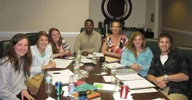 Group of people seated at a table in a meeting room. They are reviewing papers and smiling.