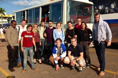 Group of diverse people standing near a bus in an outdoor setting, some smiling, some squatting.