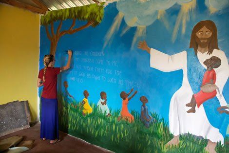 Woman painting a mural of Jesus holding a child, surrounded by other children. Blue, green, and white colors.