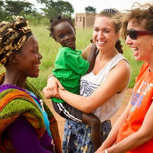 Woman holds smiling child, laughing with two other women in an outdoor setting.