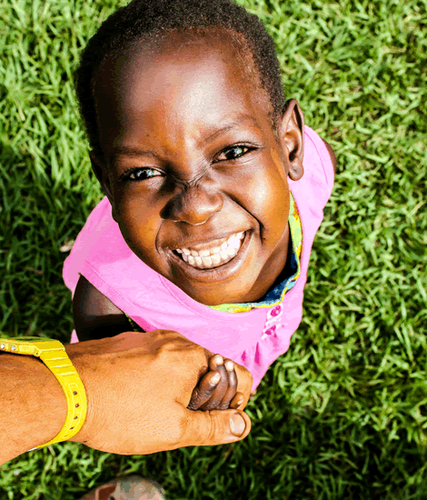 Young child with dark skin, smiling widely, holding a hand, wearing pink on green grass.