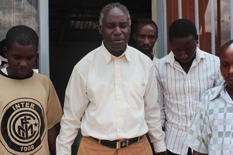 Men stand solemnly outside a building. The central figure is an older man in a light shirt; others wear shirts and a t-shirt.