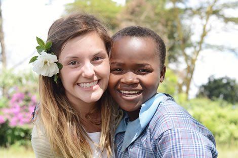 Woman with flower in her hair smiles alongside a smiling child in a school uniform outdoors.