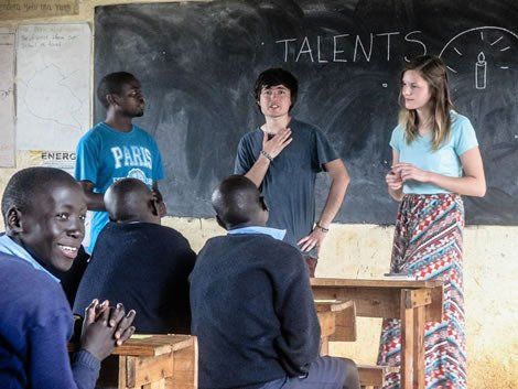 Students and teacher in a classroom. Asian male speaks; two students and a female teacher stand by chalkboard; others sit and watch.