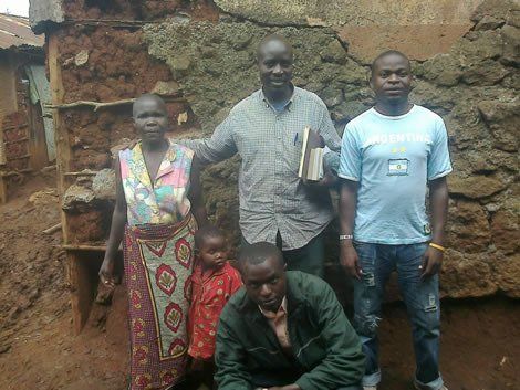 A group of five people pose outside a mud building. One man holds a book, others look at the camera.