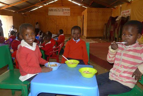 Children in red sweaters eating from bowls at a blue table in a school setting.