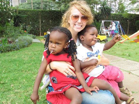 Woman in sunglasses sits with two young children in a park; one child has a lollipop.