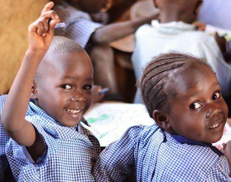 Two smiling African children in blue school uniforms, one raising hand.