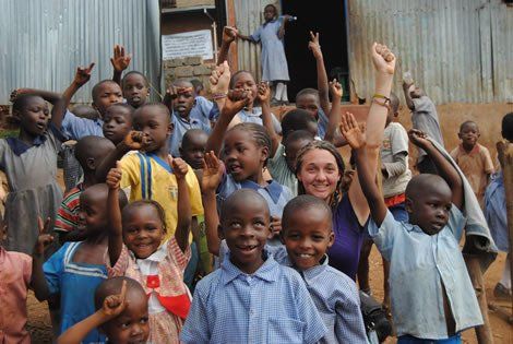 Group of children and a woman raising hands and smiling in front of a building, presumably a school.